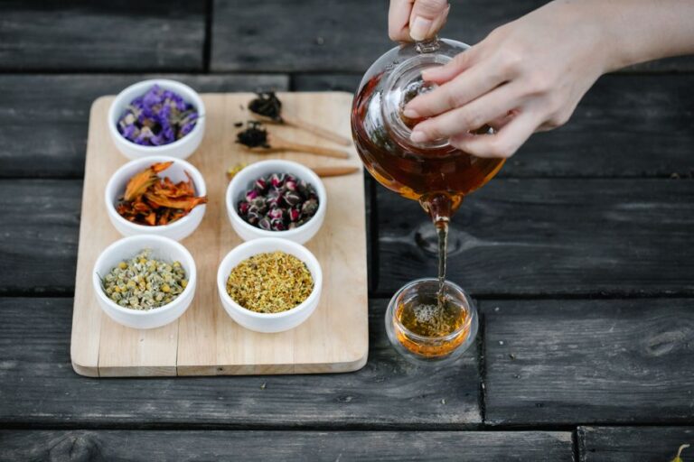 A person pours steaming herbal tea from a ceramic teapot beside dried herbs and a cup.